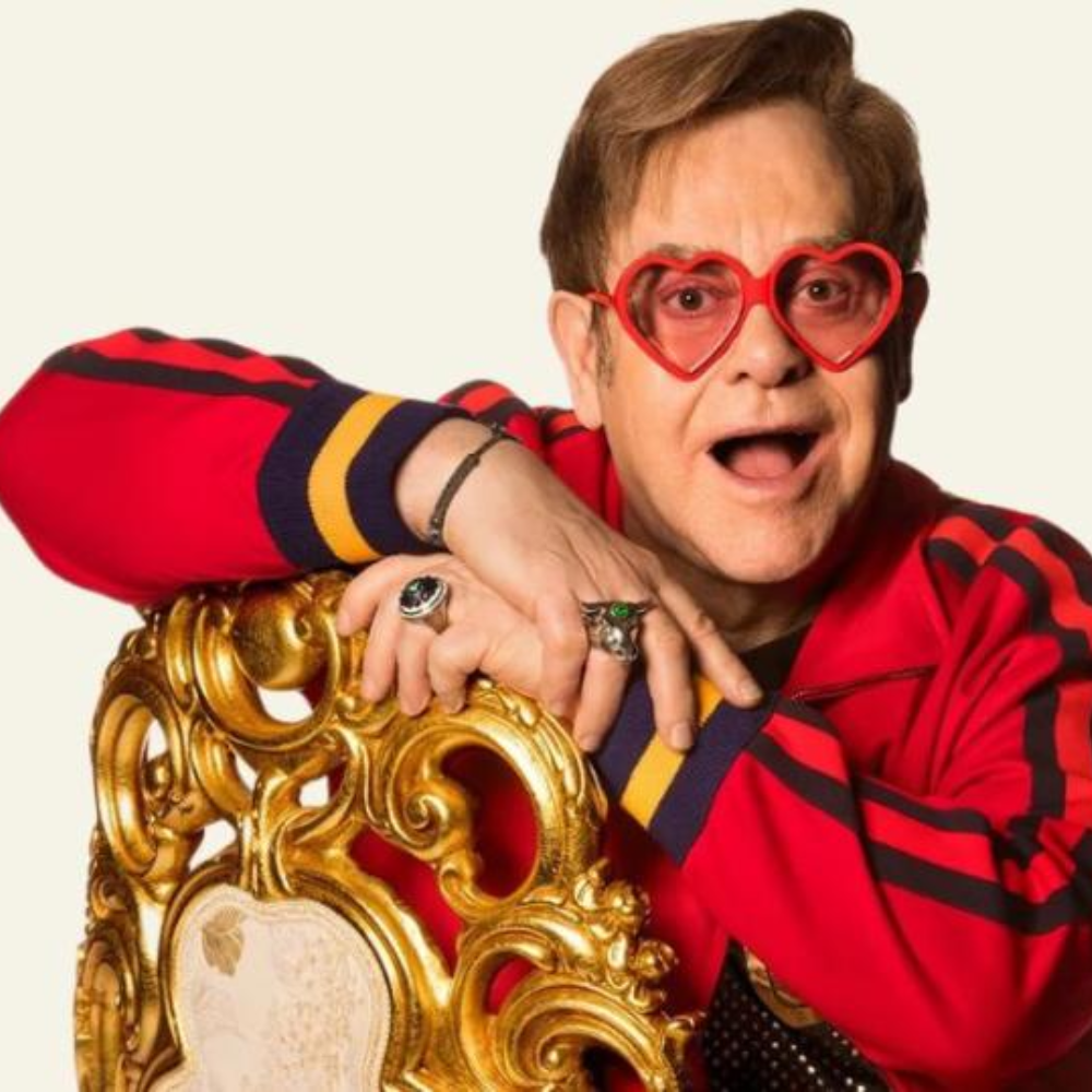 elton john sentado em uma cadeira dourada em frente a um fundo branco